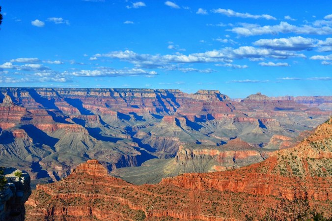 An image of the Grand Canyon which I don't even know how to describe this lol. The sky is bright blue peppered with white clouds. The canyon is colorful, similiar to a tan rainbow composed of olives, oranges, light browns, and dark browns. A handful of trees dot the bottom of the craggy canyon. What appears to be my shadow is in the bottom, left hand corner of the picture. I don't remember and only added this pic as every blog posts improves with pictures.